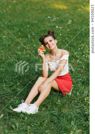 A smiling girl holds a sweet watermelon in her hands and smiles while sitting on the grass A smiling girl holds a sweet watermelon in her hands and smiles while sitting on the grass 94509825