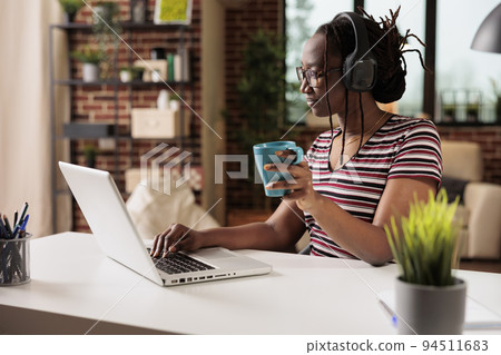Smiling woman in wireless headphones watching movie online on laptop, holding mug, drinking coffee. Indoor entertainment, leisure activity on internet, teenager in earphones relaxing Smiling woman in wireless headphones watching movie online on laptop, holding mug, drinking coffee. Indoor entertainment, leisure activity on internet, teenager in earphones relaxing 94511683