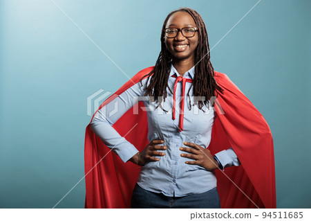 Proud superhero woman with superpower abilities wearing mighty hero cloak while smiling confident at camera. Joyful happy smiling young adult person posing as justice defender on blue background. Proud superhero woman with superpower abilities wearing mighty hero cloak while smiling confident at camera. Joyful happy smiling young adult person posing as justice defender on blue background. 94511685