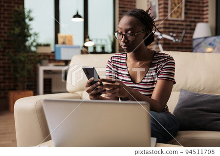 Young african american woman typing message on smartphone, browsing social media, chatting. Freelancer having break, relaxed student in glasses surfing internet on mobile phone Young african american woman typing message on smartphone, browsing social media, chatting. Freelancer having break, relaxed student in glasses surfing internet on mobile phone 94511708