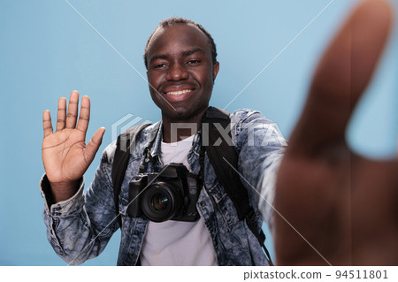 Smiling young man waving at camera while taking selfie photo on blue background. Happy photographer with DSLR device and trip backpack taking picture of himself. Studio shot 94511801