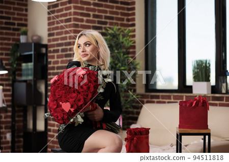 Woman posing with luxury red roses bouquet, sitting on bar stool in living room, medium shot. Beautiful blonde girl with valentines day expensive gifts, romantic date, anniversary Woman posing with luxury red roses bouquet, sitting on bar stool in living room, medium shot. Beautiful blonde girl with valentines day expensive gifts, romantic date, anniversary 94511818
