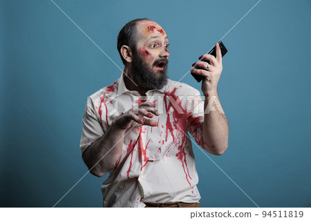 Spooky halloween monster holding smartphone in studio, using mobile phone app while being aggressive and scary. Frightening undead zombie with bloody wounds, cruel corpse with telephone. Spooky halloween monster holding smartphone in studio, using mobile phone app while being aggressive and scary. Frightening undead zombie with bloody wounds, cruel corpse with telephone. 94511819