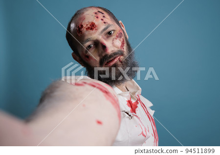 Eerie zombie holding camera to take picture, looking creepy and dangerous over blue background. Scary brain eating devil with horror look and aggressive bloody face taking frightening photo. Eerie zombie holding camera to take picture, looking creepy and dangerous over blue background. Scary brain eating devil with horror look and aggressive bloody face taking frightening photo. 94511899