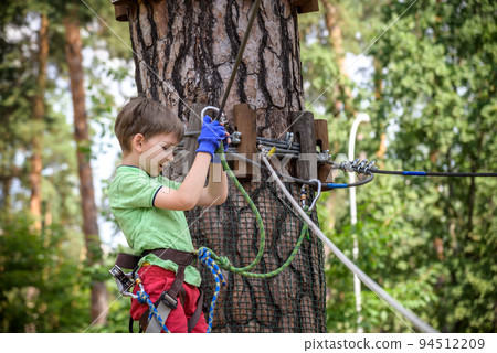 Strong excited young boy playing outdoors in rope park. Caucasian child dressed in casual clothes and sneakers at warm sunny day. Active leisure time with children concept Strong excited young boy playing outdoors in rope park. Caucasian child dressed in casual clothes and sneakers at warm sunny day. Active leisure time with children concept 94512209