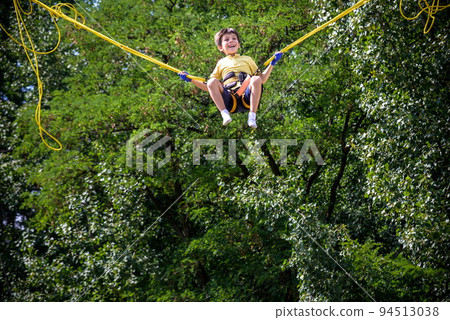 The boy is jumping on a bungee trampoline. A child with insurance and stretchable rubber bands hangs against the sky. The concept of happy childhood and games in the amusement park The boy is jumping on a bungee trampoline. A child with insurance and stretchable rubber bands hangs against the sky. The concept of happy childhood and games in the amusement park 94513038