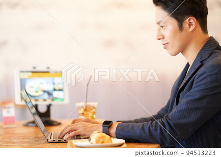 Young man using a computer while eating at a cafe 94513223