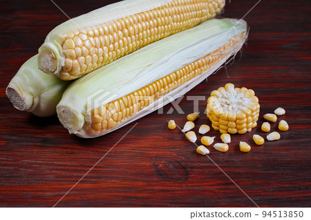 Ripe corn on the cob and a cut piece with grains around on a wooden red surface. 3 corns in half open light green wrappers, a cut disk, scattered grains of cereal on the table, space for text, closeup 94513850