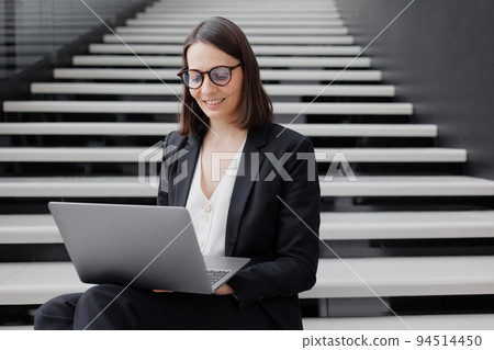business woman in a formal suit with a laptop in an office or business center sits on the steps in the lobby and works online business woman in a formal suit with a laptop in an office or business center sits on the steps in the lobby and works online 94514450