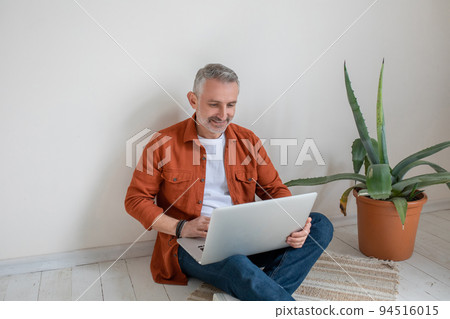 Man in terracotta shirt sitting on the floor with a laptop in hands 94516015