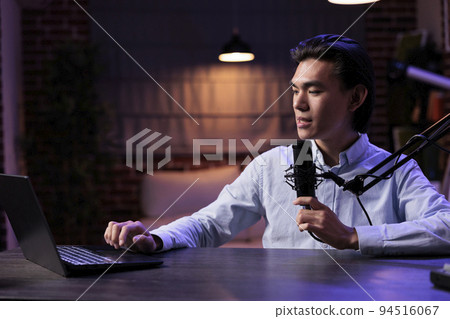 Asian vlogger recording podcast show online on social media channel, talking to audience and using laptop at sound station. Filming internet content on livestream vlog with audio production at home. 94516067