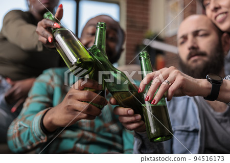 Diverse friends saying cheers and clinking beer bottles to celebrate party event at home, feeling happy about reunion. People toasting with alcohol drinks at hangout, leisure activity. Close up. 94516173