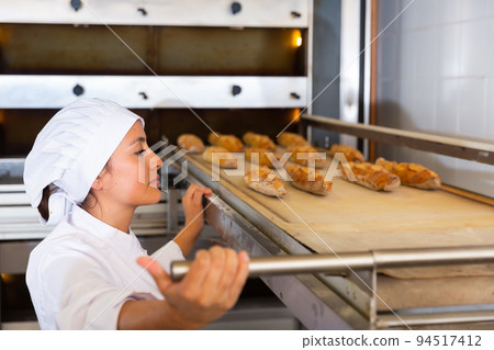 Woman pulling baked baguettes out of oven in bakery Woman pulling baked baguettes out of oven in bakery 94517412