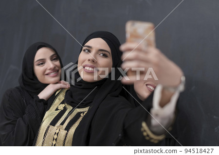 Group of young beautiful muslim women in fashionable dress with hijab using smartphone while taking selfie picture in front of black background Group of young beautiful muslim women in fashionable dress with hijab using smartphone while taking selfie picture in front of black background 94518427