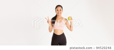 Sport, wellbeing and active lifestyle concept. Attractive asian female fitness coach, girl trainer advice eating healthy food after workout and training, standing with apple, show peace sign 94520248