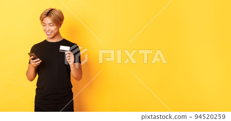 Smiling handsome asian man paying for online purchase, shopping in internet, holding smartphone and credit card, yellow background 94520259