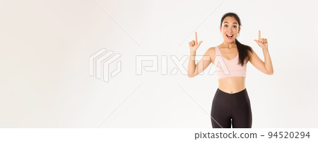 Sport, wellbeing and active lifestyle concept. Excited and happy asian fitness girl in sportswear, pointing fingers up and looking amazed with great discounts, read banner, white background Sport, wellbeing and active lifestyle concept. Excited and happy asian fitness girl in sportswear, pointing fingers up and looking amazed with great discounts, read banner, white background 94520294