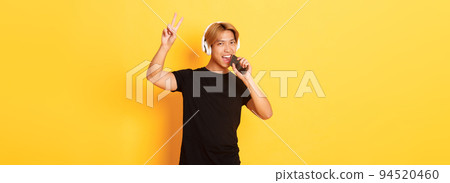 Sassy handsome asian guy in headphones, playing karaoke app, singing into mobile phone microphone, showing peace gesture, standing yellow background Sassy handsome asian guy in headphones, playing karaoke app, singing into mobile phone microphone, showing peace gesture, standing yellow background 94520460