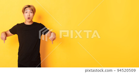 Amused happy asian guy, pointing fingers down and gasping impressed, showing awesome advertisement, yellow background Amused happy asian guy, pointing fingers down and gasping impressed, showing awesome advertisement, yellow background 94520509