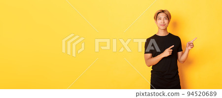 Smiling asian male student with blond hair, pointing fingers right, showing logo, standing yellow background Smiling asian male student with blond hair, pointing fingers right, showing logo, standing yellow background 94520689