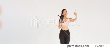 Full length of smiling slim and healthy asian girl taking selfie fitness training session, female blogger and athelte showing water bottle on camera of mobile phone, standing over white background Full length of smiling slim and healthy asian girl taking selfie fitness training session, female blogger and athelte showing water bottle on camera of mobile phone, standing over white background 94520999