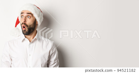 Close-up of amazed bearded man in santa hat, looking left with surprised face, christmas promo offer, standing over white background 94521182