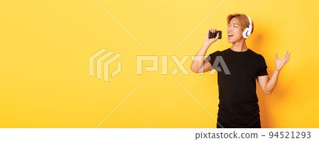 Joyful attractive korean guy in headphones, playing karaoke app, singing into mobile phone microphone, standing yellow background Joyful attractive korean guy in headphones, playing karaoke app, singing into mobile phone microphone, standing yellow background 94521293