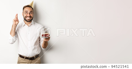 Holidays and celebration. Happy man having birthday party, making wish on b-day cake and cross fingers for good luck, standing against white background Holidays and celebration. Happy man having birthday party, making wish on b-day cake and cross fingers for good luck, standing against white background 94521541