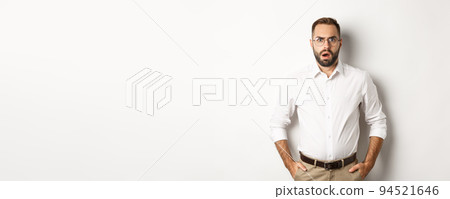 Shocked and displeased businessman in glasses, gasping and looking upset at camera, standing over white background 94521646