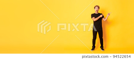 Full-length of amazed and impressed attractive asian guy pointing fingers upper right corner, showing logo, yellow background 94522654
