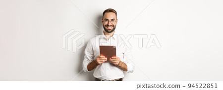 Handsome bearded man in glasses using digital tablet, smiling satisfied, standing against white background 94522851