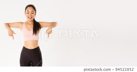 Sport, wellbeing and active lifestyle concept. Excited and happy asian fitness girl in sportswear, pointing fingers down and looking amazed with great discounts, read banner, white background 94522852