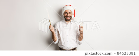Winter holidays and celebration. Handsome bearded man having New Year party, holding firework sparkle and champagne, wearing santa hat, white background 94523391