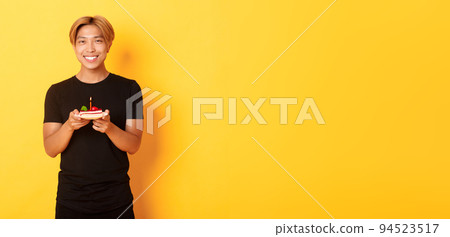 Handsome happy asian blond guy, smiling pleased as celebrating birthday, holding b-day cake, standing over yellow background 94523517
