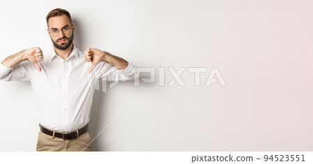 Displeased businessman in glasses showing thumbs down, dislike and disapprove, standing over white background 94523551