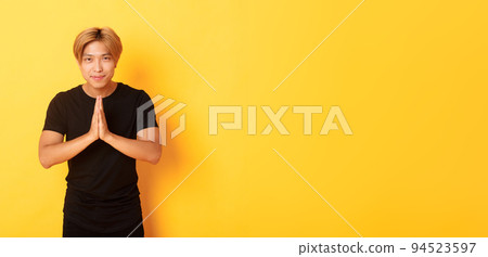 Portrait of friendly and polite asian blond guy, clasp hands together in namaste gesture, bowing and greeting person, standing over yellow background 94523597