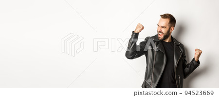 Confident bearded man celebrating victory, winning prize, making fist pump and rejoicing, wearing black leather jacket, white background 94523669