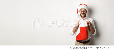Happy adult man open christmas sock and looking inside, receiving xmas gifts for winter holidays, standing in santa hat, white background 94523858