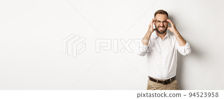 Businessman having headache, grimacing and holding hands on head, standing over white background Businessman having headache, grimacing and holding hands on head, standing over white background 94523958
