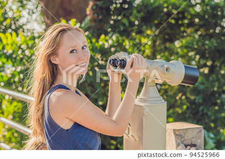 young beautiful blond woman enjoy the view with an coin operated binoculars. The water and the sky is blue. she wears a white dress and sunglasses. she feels good, is smiling and look to the horizon 94525966