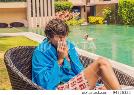 Sick man traveler. The man caught a cold on vacation, sits sad at the pool drinking tea and blows his nose into a napkin 94526323