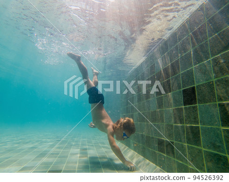 A child boy is swimming underwater in a pool, smiling and holding breath, with swimming glasses 94526392