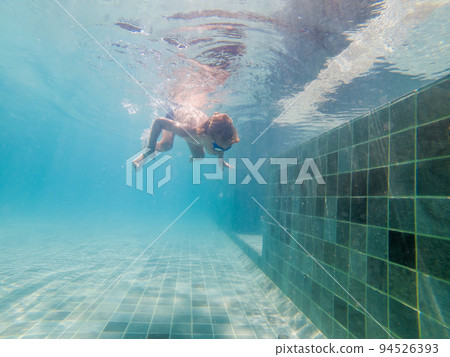 A child boy is swimming underwater in a pool, smiling and holding breath, with swimming glasses 94526393