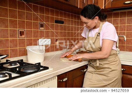 Beautiful brunette woman, pleasant housewife in beige chef's apron standing by a kitchen countertop and using a rolling pin, rolls out dough for bread, pizza, pie or a homemade festive tartlets 94527059