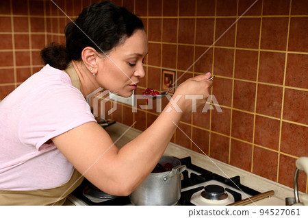 Close-up of a middle-aged multi-ethnic pretty woman, a housewife tasting caramelized cherries while cooking in the home kitchen 94527061