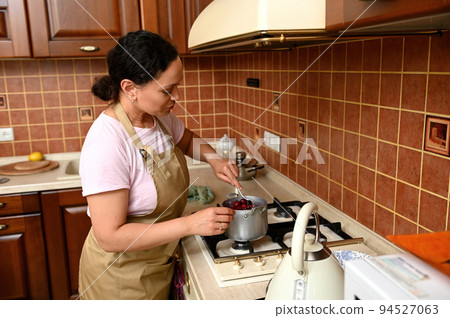 Beautiful multiethnic young brunette woman, a pleasant housewife, in a beige chef's apron, stands at the stove in the home kitchen and cooks caramelized cherries in a saucepan for a festive pie. 94527063