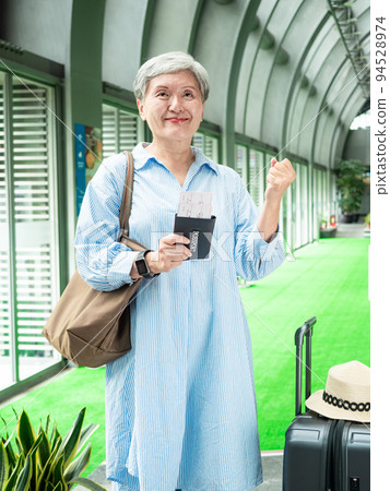 60多歲的亞洲熟年婦女微笑著站著,身旁有手提箱行李袋和手持登機證與護照,適用於covid19疫情後旅 60多歲的亞洲熟年婦女微笑著站著,身旁有手提箱行李袋和手持登機證與護照,適用於covid19疫情後旅 94528974