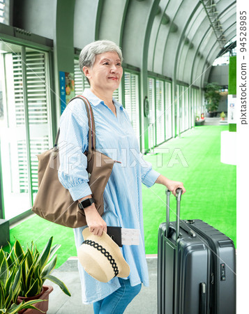 60多歲的亞洲熟年婦女微笑著站著，身旁有手提箱行李袋和手持登機證與護照，適用於covid19疫情後旅 94528985