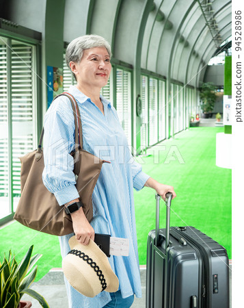 Asian mature woman in her 60s smiling and standing with suitcase duffel bag and holding boarding pass and passport for travel after covid19 94528986
