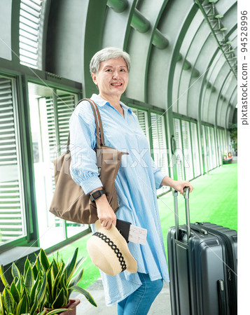 Asian mature woman in her 60s smiling and standing with suitcase duffel bag and holding boarding pass and passport for travel after covid19 Asian mature woman in her 60s smiling and standing with suitcase duffel bag and holding boarding pass and passport for travel after covid19 94528996
